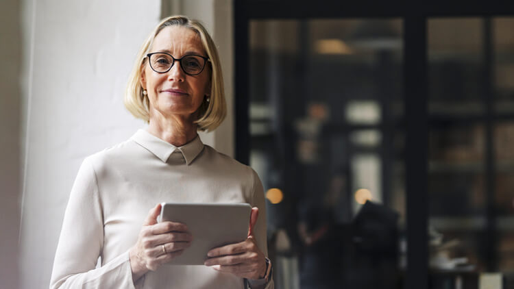 Older business woman stood next to a window in an office holding a tablet device looking into the camera