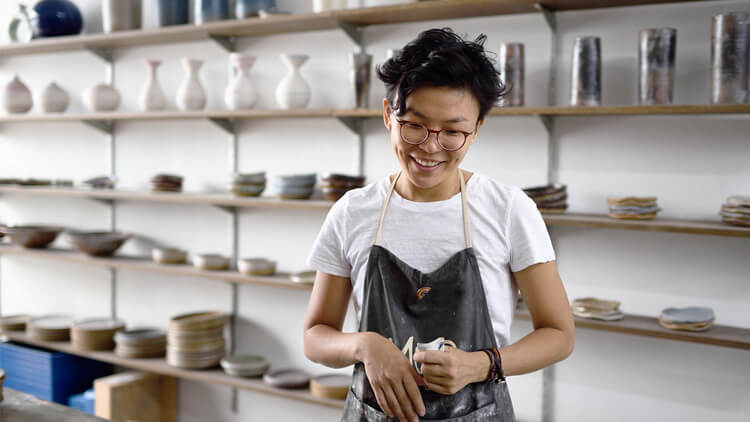 Asian woman stood in her pottery workshop looking down admiring her creation 